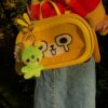 Person wearing rainbow striped sweate, a jean skirt with a Yellow cross body dog bag with an smiling alien plush keychain hanging off of the bag zipper