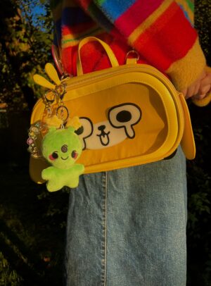 Person wearing rainbow striped sweate, a jean skirt with a Yellow cross body dog bag with an smiling alien plush keychain hanging off of the bag zipper