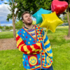 Man standing in a green field holding balloons while wearing a primary striped tee, jeans and a red, yellow and blue cardigan.