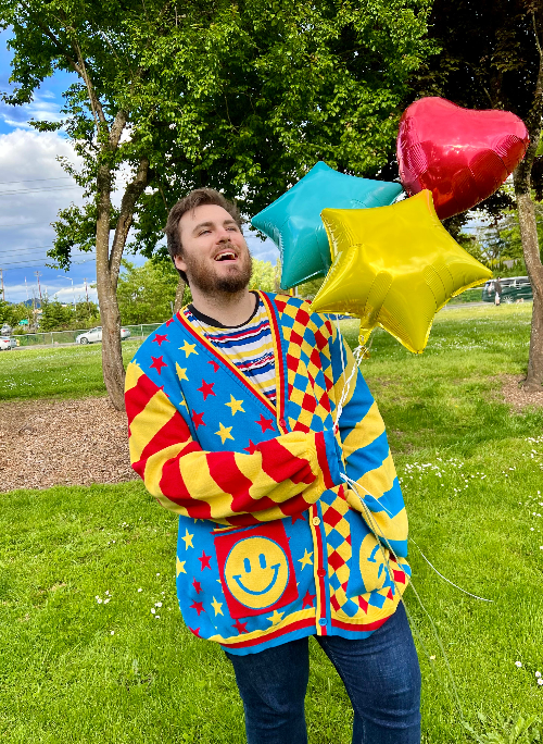 Man standing in a green field holding balloons while wearing a primary striped tee, jeans and a red, yellow and blue cardigan.