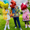 Two women and one one man standing in a green field holding balloons that cover their faces. One girl is wearing a yellow egg cardigan, blue shirt, white skirt and a fanny pack. Man standing in the center is wearing jeans and a primary colored cardigan. Last woman is wearing a pink capsule and heart patterned cardigan, a black and pink dress and legwarmers.
