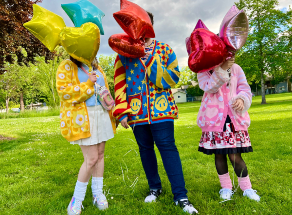 Two women and one one man standing in a green field holding balloons that cover their faces. One girl is wearing a yellow egg cardigan, blue shirt, white skirt and a fanny pack. Man standing in the center is wearing jeans and a primary colored cardigan. Last woman is wearing a pink capsule and heart patterned cardigan, a black and pink dress and legwarmers.