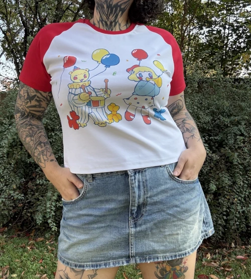 A woman wearing a white crop top with red short sleeves and a short jean skirt standing outside in front of a tree. The shirt has a white body with playful primary colored clown characters, balloons and confetti printed on it.