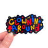 A hand holding a sticker with the text 'CLOWNS AROUND' in a colorful font