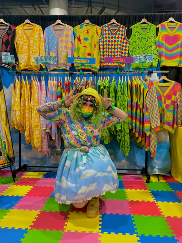 Girl on one knee bending down on colorful mats in front of various clothing racks with colorful cardigans. she is wearing a yellow bucket hat, a rainbow mask, a rainbow button up shirt over a dress with a sky