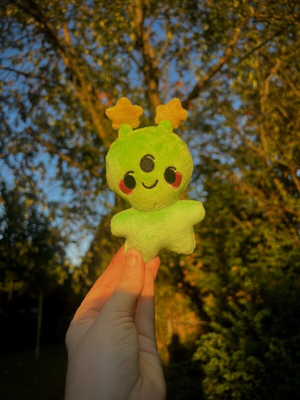 Smiling green alien plush held in front of a tree with a blurred background