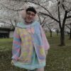 A person wearing a multicolored pastel clown patterned cardigan, paired with a collared blouse and striped skirt, standing outdoors.