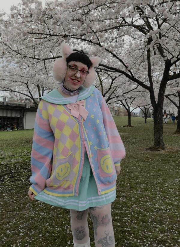 A person wearing a multicolored pastel clown patterned cardigan, paired with a collared blouse and striped skirt, standing outdoors.