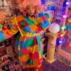 Girl is standing facing away from a mirror and looking down at the ground. She is wearing a knit sweater dress with a colorful puzzle playmat design. she is also wearing a yellow belt, pink hair, and red bear slippers. Behind her is a collage of miscellaneous retro book covers.