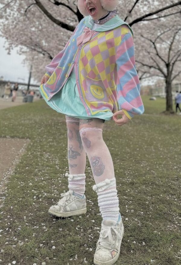A person wearing a multicolored pastel clown patterned cardigan, paired with a collared blouse and striped skirt, standing outdoors.
