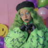 Girl with green hair standing in front of a pink background wearing a black beret, black gloves and a green cardigan with a slime pattern.