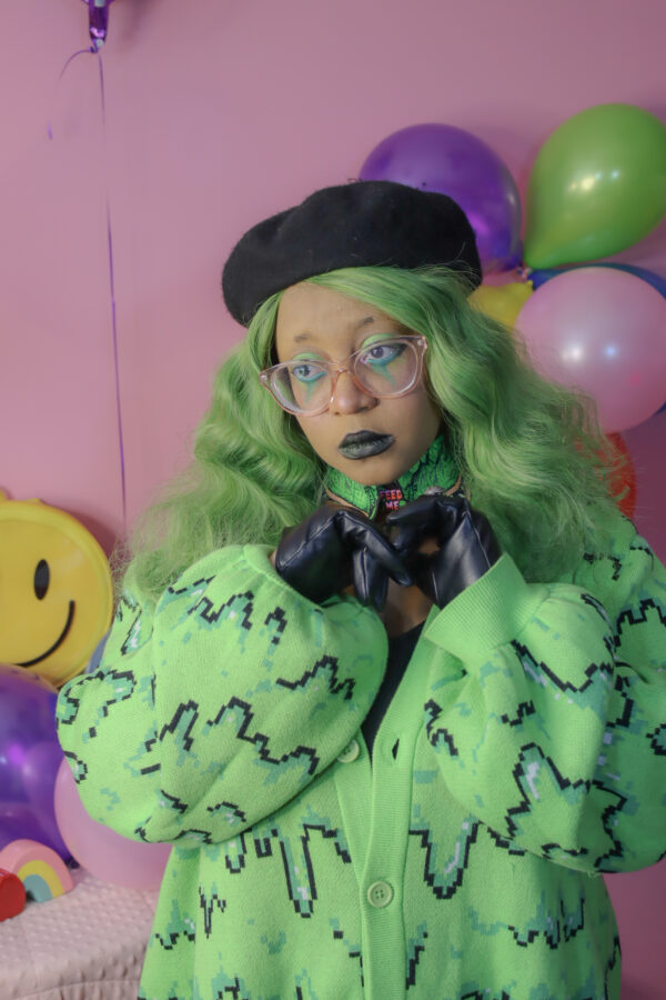 Girl with green hair standing in front of a pink background wearing a black beret, black gloves and a green cardigan with a slime pattern.