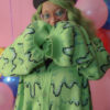 Girl with green hair standing in front of a pink background with rainbow balloons wearing a black beret, black gloves and a green cardigan with a slime pattern.
