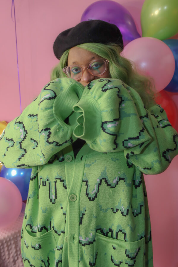 Girl with green hair standing in front of a pink background with rainbow balloons wearing a black beret, black gloves and a green cardigan with a slime pattern.