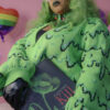 Girl with green hair standing in front of a pink background holding a "Plants that Kill" book wearing a black beret, black gloves and a green cardigan with a slime pattern.