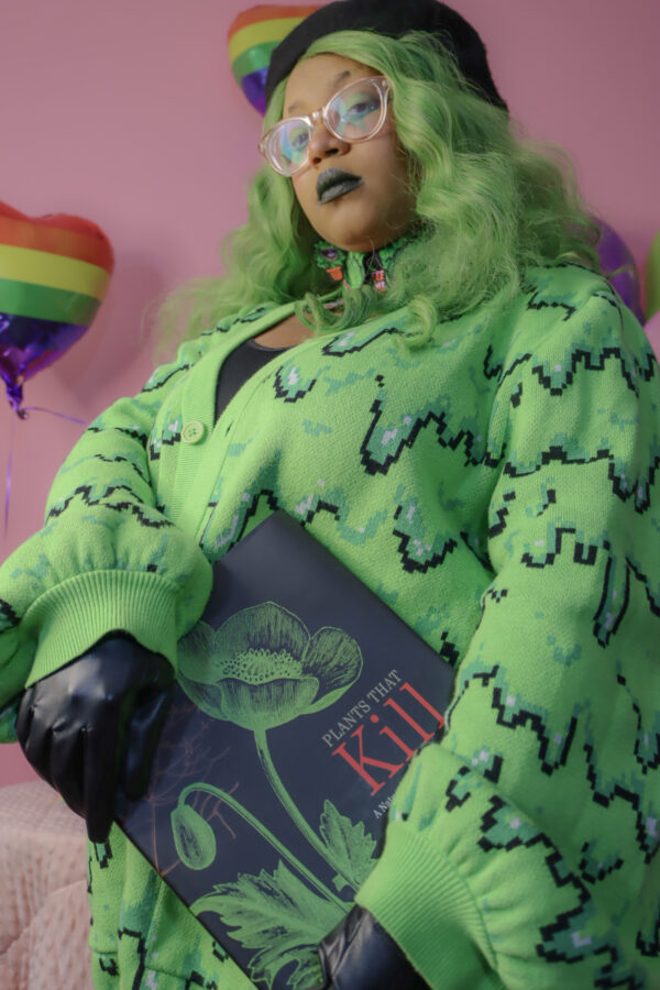 Girl with green hair standing in front of a pink background holding a "Plants that Kill" book wearing a black beret, black gloves and a green cardigan with a slime pattern.