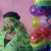 Girl with green hair standing in front of a pink background holding a rainbow heart balloon wearing a black beret, black gloves and a green cardigan with a slime pattern.