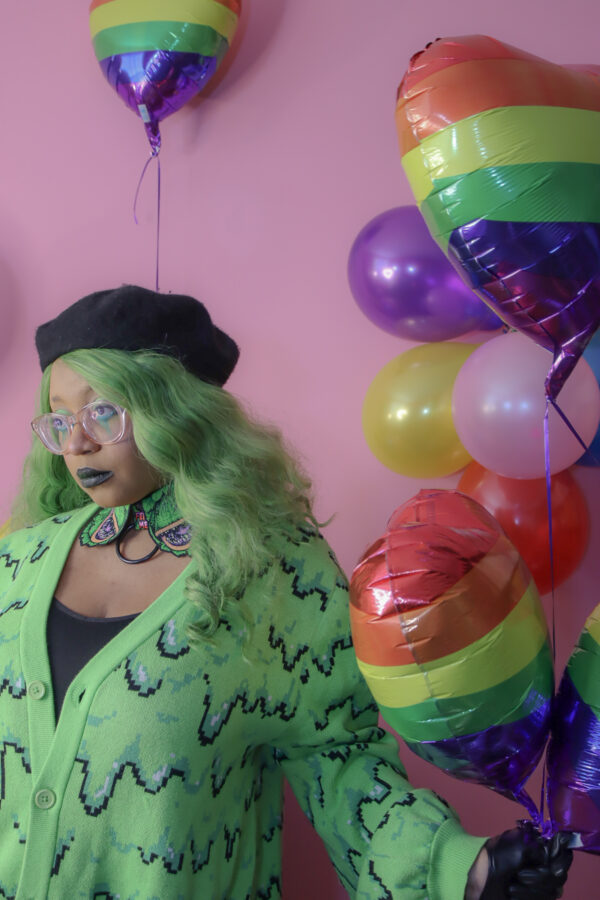 Girl with green hair standing in front of a pink background holding a rainbow heart balloon wearing a black beret, black gloves and a green cardigan with a slime pattern.