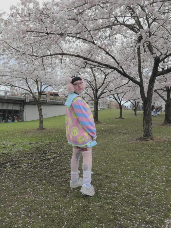 A person wearing a multicolored pastel clown patterned cardigan, paired with a collared blouse and striped skirt, standing outdoors.