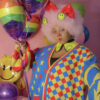 Woman wearing clown makeup, looking at the camera with one hand on her face and the other holding a few rainbow balloons. She is in front a pink background wearing glasses, a happy face collar, a big red bow and a red, blue and yellow cardigan with stars and diamond patterns on it.