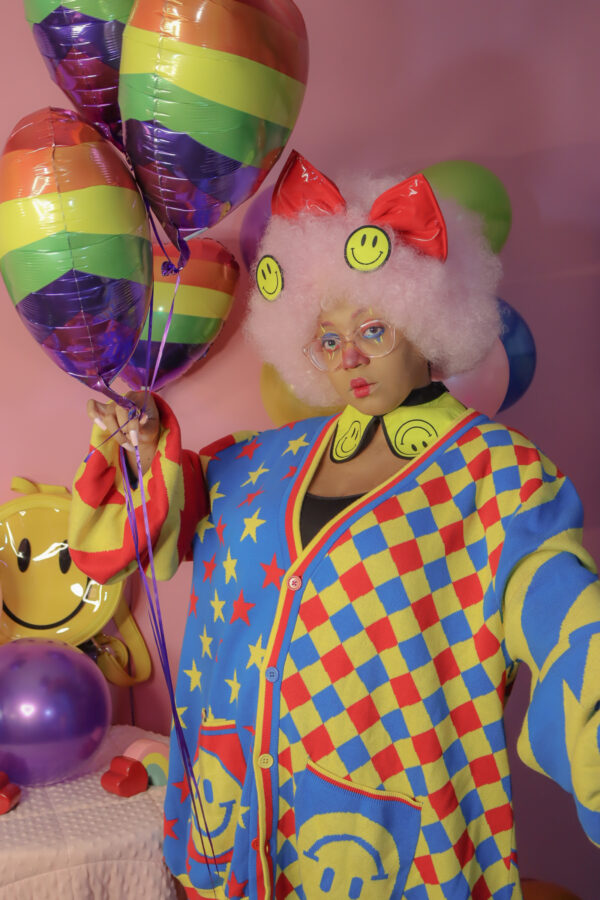 Woman wearing clown makeup, looking at the camera with one hand on her face and the other holding a few rainbow balloons. She is in front a pink background wearing glasses, a happy face collar, a big red bow and a red, blue and yellow cardigan with stars and diamond patterns on it.