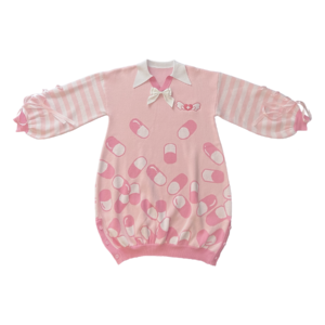 Flat lay image of the front of the pink menhera sweater dress. The sweater dress body is pastel pink with darker pink and white capsules on it. It has a white pointed collar and a removable white ribbon on the center collar. The sleeves are pastel pink and white striped with pastel pink ribbons installed to form a bow on the end of the sleeves. There is a darker pink and white embroidered heart with a cross and wings on the front left chest.