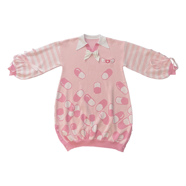 Flat lay image of the front of the pink menhera sweater dress. The sweater dress body is pastel pink with darker pink and white capsules on it. It has a white pointed collar and a removable white ribbon on the center collar. The sleeves are pastel pink and white striped with pastel pink ribbons installed to form a bow on the end of the sleeves. There is a darker pink and white embroidered heart with a cross and wings on the front left chest.