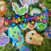 hand holding a colorful sticker in front of other sticker designs on grass. The sticker being held is colorful and says "understimulated" sticker with confetti and stars in the background.