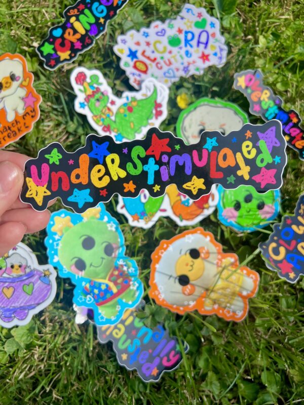 hand holding a colorful sticker in front of other sticker designs on grass. The sticker being held is colorful and says "understimulated" sticker with confetti and stars in the background.