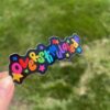 A hand holding a colorful sticker with the text 'overstimulated' in a decorative font, against a blurred background of green grass.