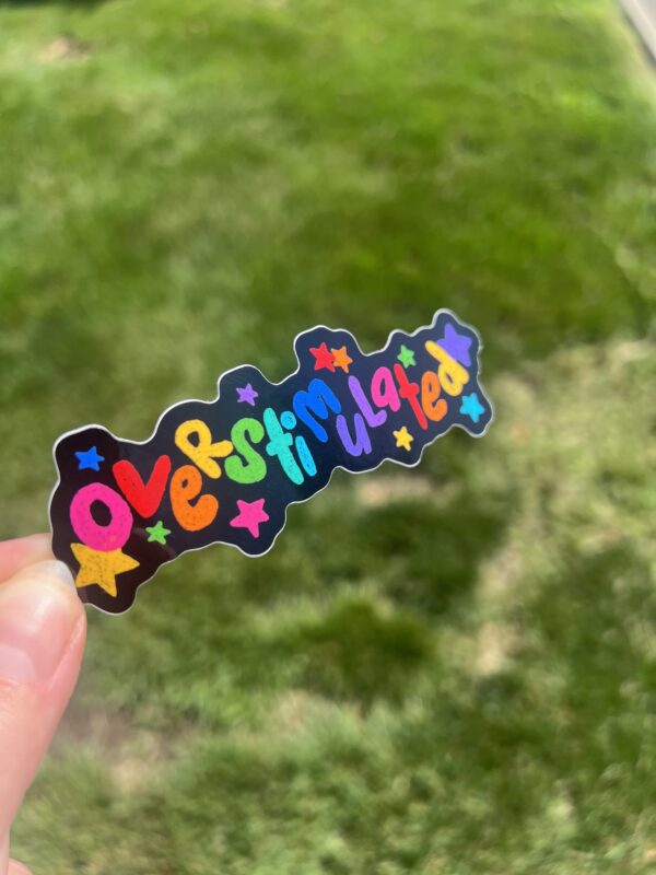 A hand holding a colorful sticker with the text 'overstimulated' in a decorative font, against a blurred background of green grass.