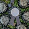Black rosette pins with the text 'Not Cosplay' printed in silver, laying on grass. One rosette is facing down wards with its pin facing up to the camera.