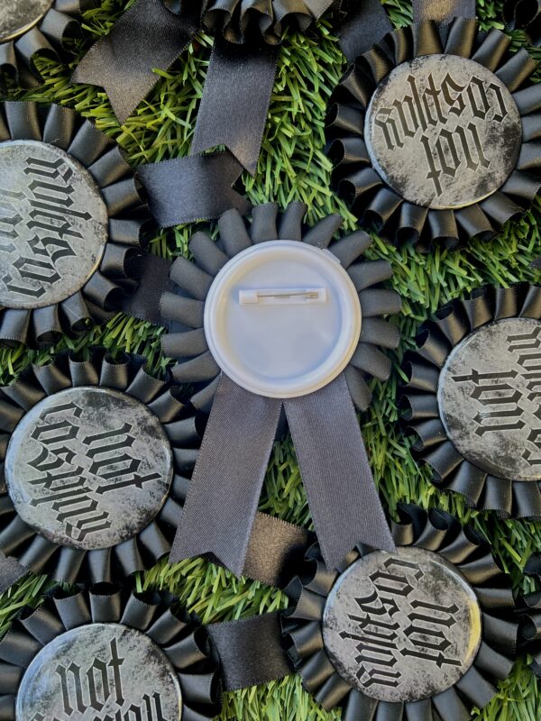 Black rosette pins with the text 'Not Cosplay' printed in silver, laying on grass. One rosette is facing down wards with its pin facing up to the camera.