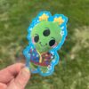 An alien plush sticker with a cartoon character design, held up against an outdoor background.