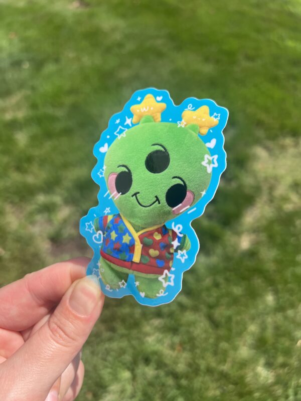 An alien plush sticker with a cartoon character design, held up against an outdoor background.