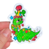 Hand holding a sticker of a Green dinosaur on a clear background. Green Dinosaur is wearing a party hat on its head and a colorful bow tie.