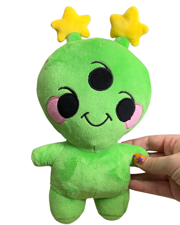 hand holding medium sized cheerful alien plush. Alien has three eyes, pink round cheeks, two yellow star antennae
