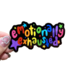 A sticker with the phrase 'emotionally exhausted' written in a colorful, playful font, held between fingers