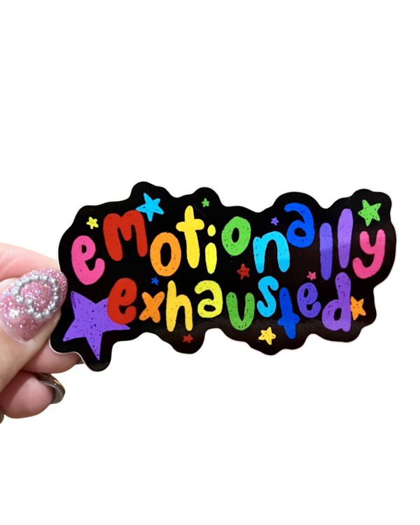 A sticker with the phrase 'emotionally exhausted' written in a colorful, playful font, held between fingers