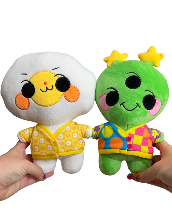 Two hands holding two different plush in each hand facing the camera. Hand on the right is holding a cheerful alien plushie wearing a rainbow cardigan. And the hand on the left is holding an mischievous alien plush wearing a yellow and orange cardigan.