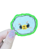 A hand holding a round egg sticker with a character illustration on it on a clear backdrop.