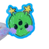 A sticker with a cheerful green alien character design being held by a persons hand on a clear background