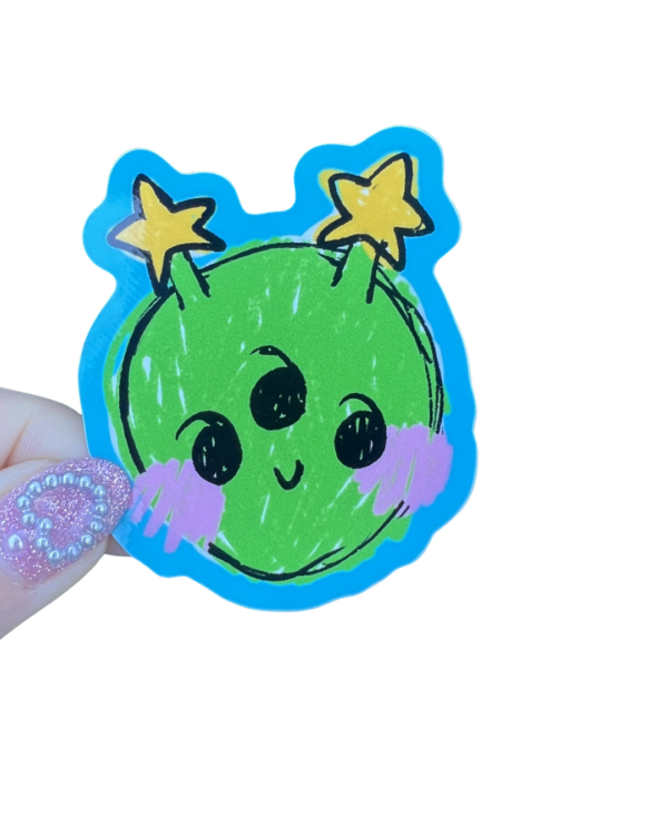 A sticker with a cheerful green alien character design being held by a persons hand on a clear background