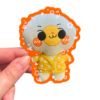 A plush sticker of a cartoon character with a yellow outfit and orange accents, being held up against a clear background
