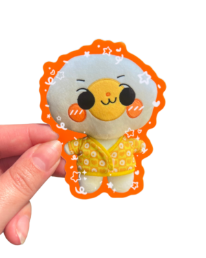 A plush sticker of a cartoon character with a yellow outfit and orange accents, being held up against a clear background