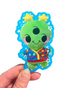 A plush alien sticker with a cartoon character design, held up against a clear background