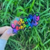 Suggestion A sticker with the phrase 'emotionally exhausted' written in a colorful, playful font, held between fingers against a grassy background.