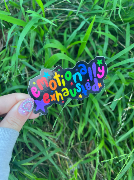 Suggestion A sticker with the phrase 'emotionally exhausted' written in a colorful, playful font, held between fingers against a grassy background.