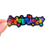 A hand holding a colorful sticker with the text 'overstimulated' in a decorative font and confetti on a clear background