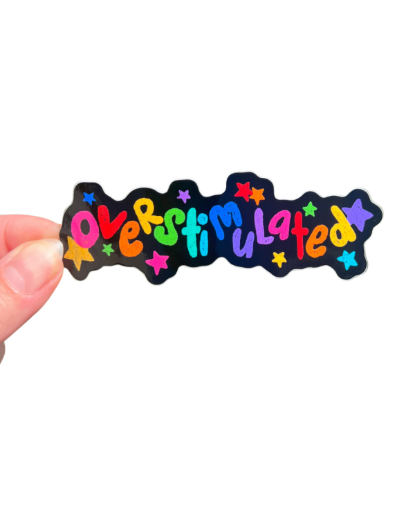A hand holding a colorful sticker with the text 'overstimulated' in a decorative font and confetti on a clear background